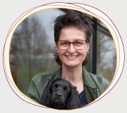Rabea Wienholt, die einen schwarzen Hund mit langen Ohren auf den Armen hält, draußen vor einem Fenster stehend, mit Bäumen im Hintergrund.
