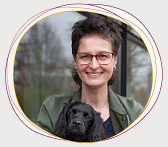 Rabea Wienholt, die einen schwarzen Hund mit langen Ohren auf den Armen hält, draußen vor einem Fenster stehend, mit Bäumen im Hintergrund.