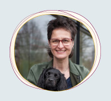 Rabea Wienholt, die einen schwarzen Hund mit langen Ohren auf den Armen hält, draußen vor einem Fenster stehend, mit Bäumen im Hintergrund.